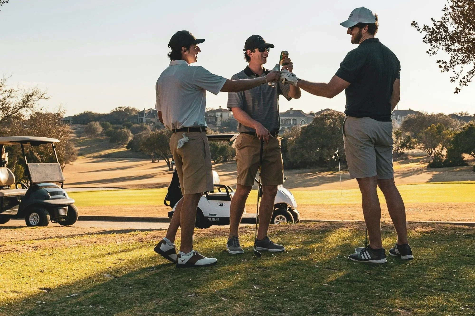 Three men on a golf course exchanging a handshake, with golf carts and trees in the background during sunset.